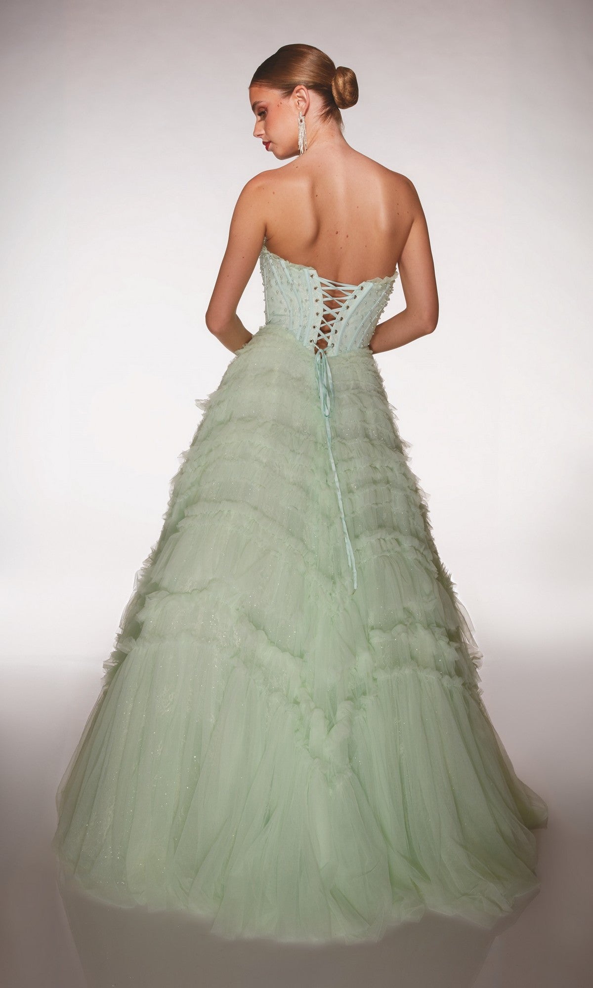A woman with blonde hair in a bun models the Alyce 62208 long prom dress, a pale green strapless ballgown with a lace-up back and tulle ruffle skirt, shown from behind against a plain background.